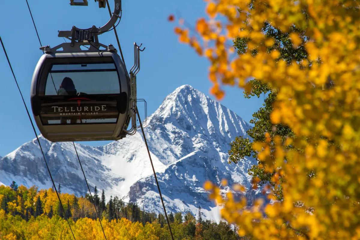 A scenic exterior shot featuring a gondola ascending towards a snow-capped mountain, with vibrant autumn foliage in the foreground. The image captures the connection between the town and the rugged natural environment, highlighting year-round adventure possibilities.