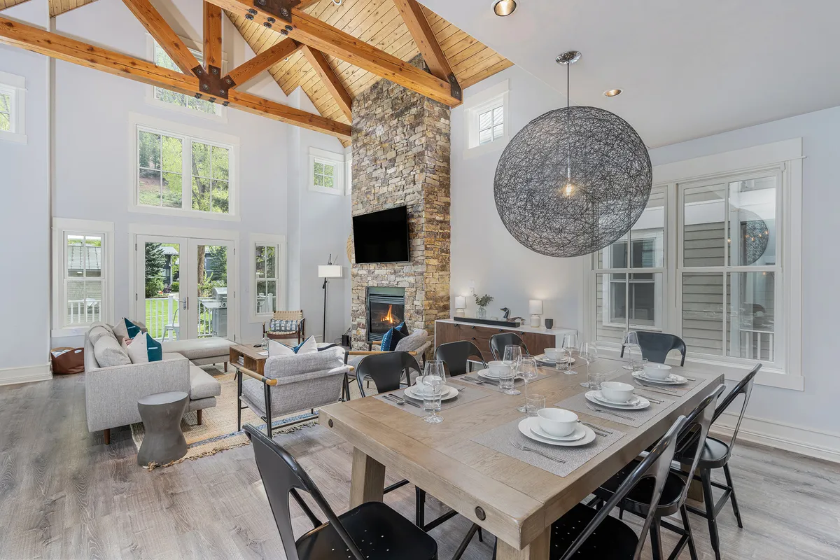 This interior shot showcases the large stone fireplace as a focal point, a dining table set for a meal, expansive windows letting in natural light, exposed wooden beams, and a modern pendant light. The scene is warm and inviting, filled with the sense of family gatherings and socializing in a luminous open space.
