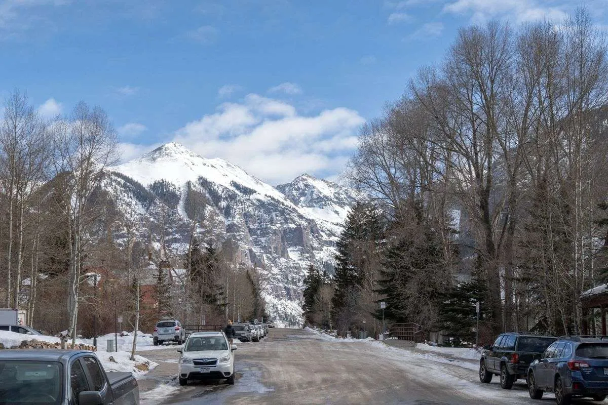 A scenic, tree-lined street set against snow-capped mountains, with parked cars and glimpses of the sky creating a serene, inspiring atmosphere. The image conveys easy access to nature, mountain vistas, and neighborhood tranquility.