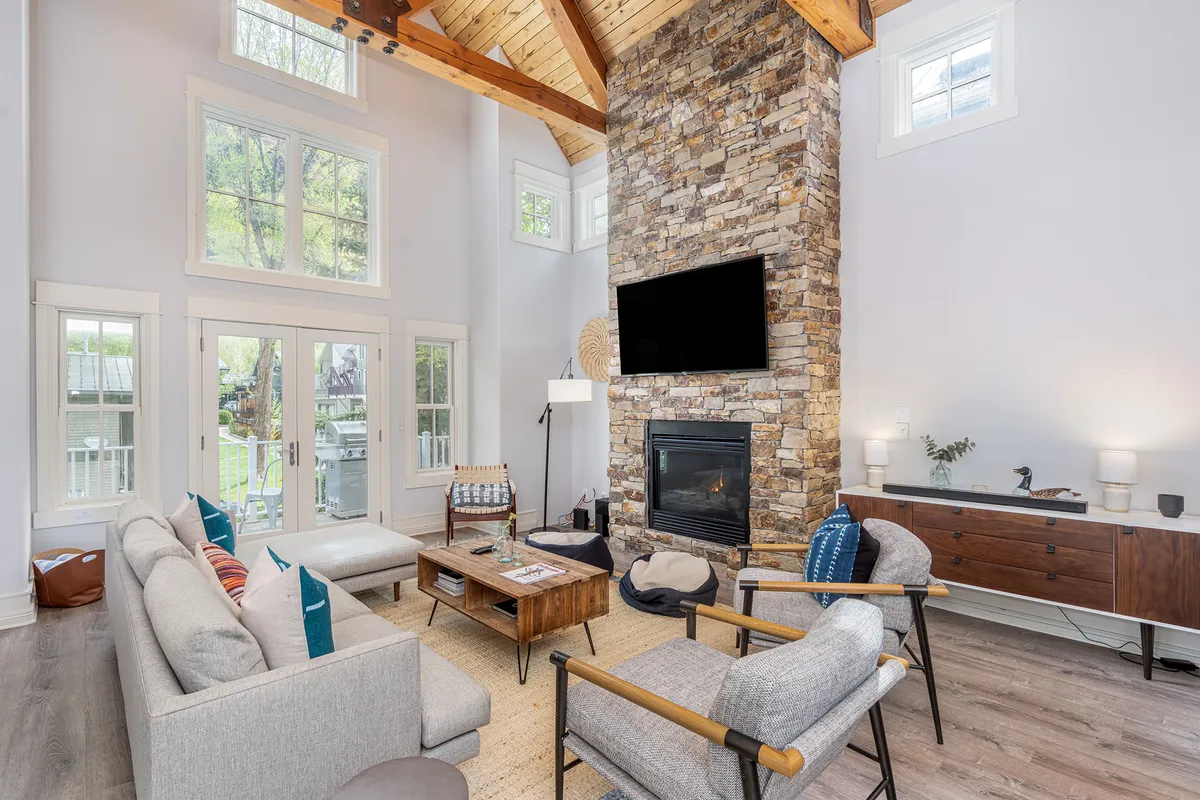 The image showcases a spacious, sun-drenched living area featuring striking wooden beams, a grand stone fireplace, floor-to-ceiling windows with sweeping mountain views, and inviting modern furnishings arranged for comfort and conversation.