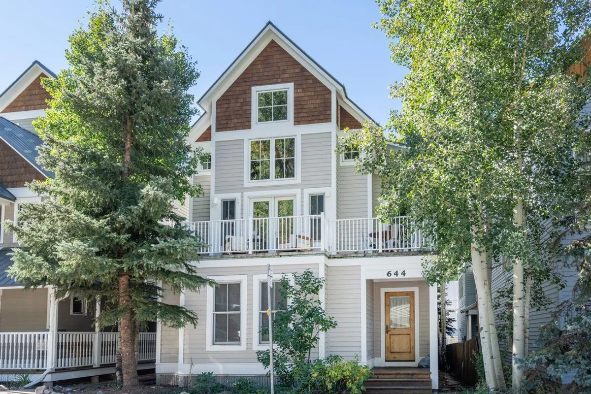 The front porch with railing, large windows, and a wooden door, framed by surrounding trees. The setting evokes a cozy, welcoming vibe and the promise of sunny afternoons or starlit nights spent relaxing or entertaining guests outdoors.