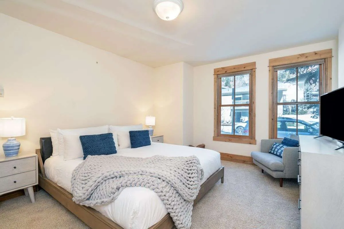 This image features a large bed with crisp white bedding, a textured gray throw blanket, wooden framed windows letting in ample natural light, and nightstands with lamps. The atmosphere is warm, inviting, and restful—ideal for showcasing a serene bedroom retreat.