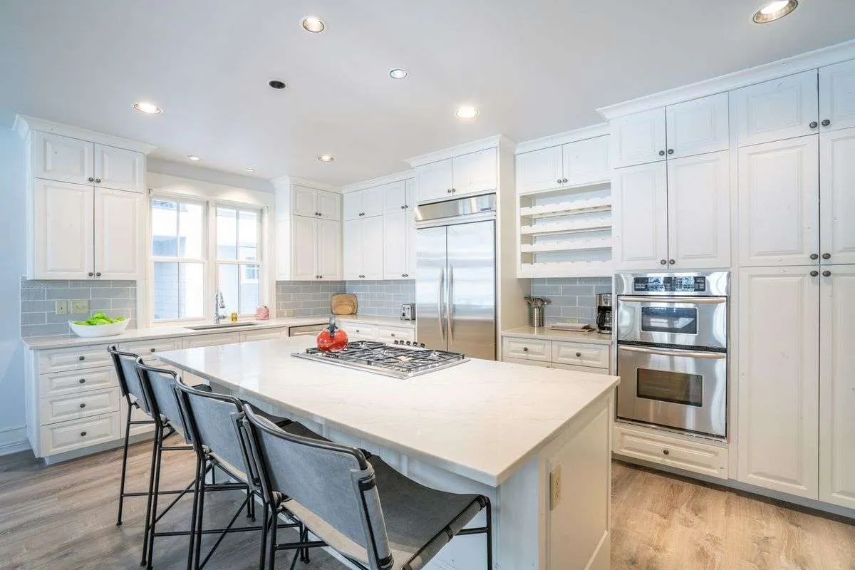 A view of the gourmet kitchen with a spacious island, stainless steel appliances, and white cabinetry. The scene exudes modern comfort and functionality, suggesting meal preparations, cooking together, and joyful gatherings.