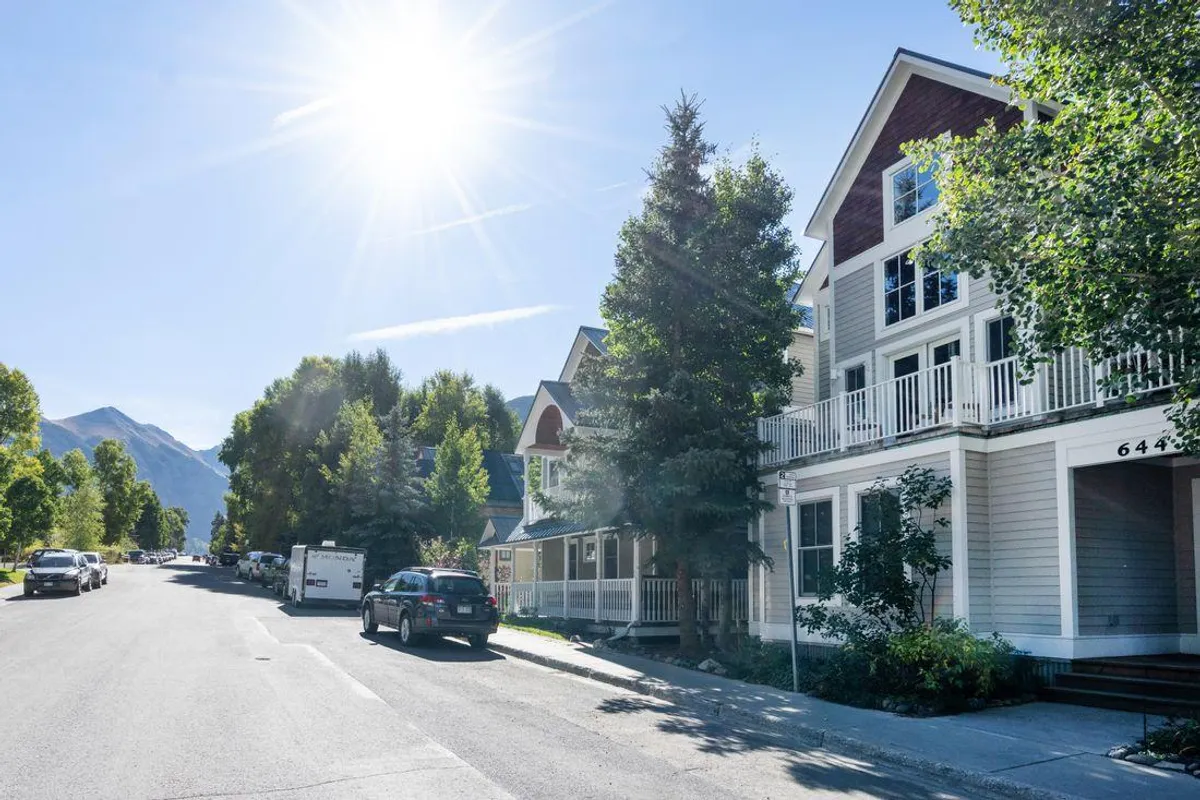 A cheerful exterior street scene with sunlight streaming down on a charming, tree-lined neighborhood. The houses display distinctive architectural details, and people can be imagined walking or biking along the street, enjoying the relaxed, welcoming atmosphere. The mood hints at weekend barbecues and community gatherings, aligning with the idea of local festivals and neighborhood fun.