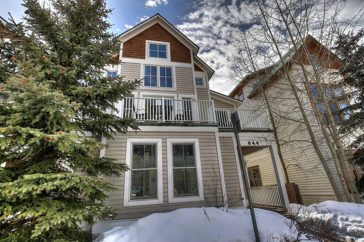 A welcoming exterior view of a three-story mountain retreat with balconies overlooking a snowy landscape and a prominent evergreen tree. The home exudes comfort and luxury amidst the serene mountain environment, suggesting a perfect place to return after a day outdoors.