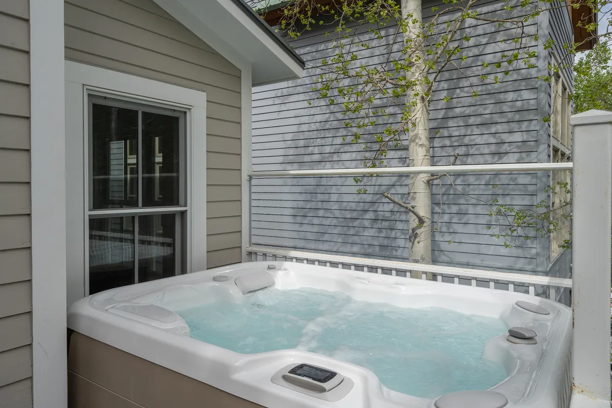 This image features the private outdoor hot tub nestled among trees, with the exterior wall of the home in view. The tranquil setting invites relaxation, with ample space for small gatherings or personal rejuvenation after an active day. The sky above hints at stargazing possibilities.