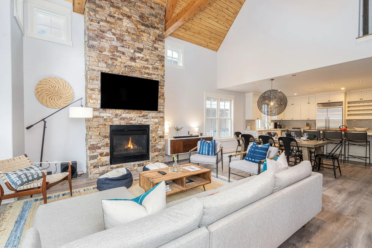 A modern, cozy living room centers on a stone fireplace, with a comfortable sofa and large windows letting in mountain light. Multiple spaces for gathering, relaxing, or enjoying quiet evenings are visible, making this a perfect spot for unwinding indoors.